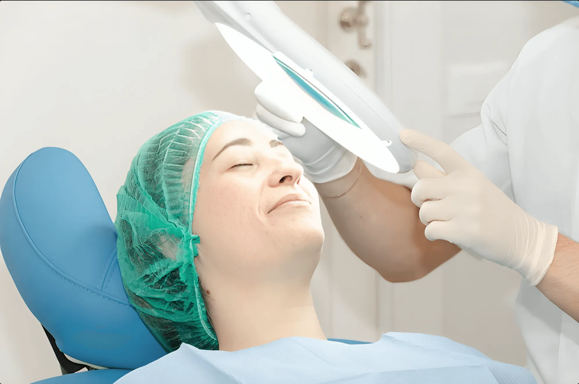 Una paziente con cuffia per capelli e camice medico si sdraia su una sedia mentre un operatore sanitario munito di guanti presso la Medicina Estetica e Dermatologia Coppede Medical Hub Roma regola una lampada da visita sopra il suo viso.