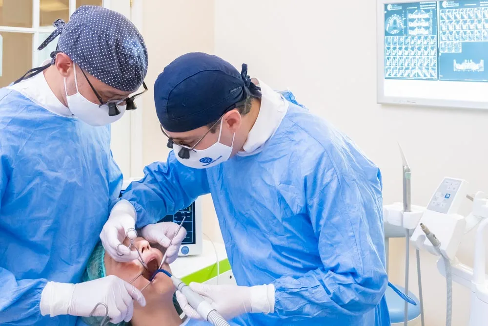 Due dentisti in camice chirurgico e mascherina eseguono una procedura odontoiatrica presso l'Implantologia-Coppede-Medical-Hub-Roma, con attrezzature mediche visibili sullo sfondo.