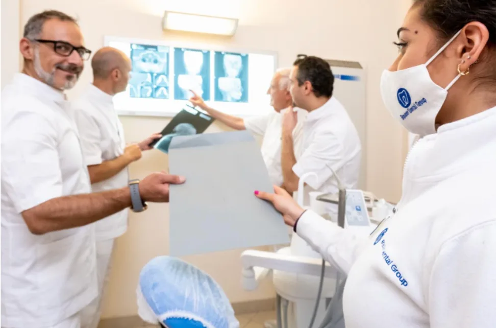 Quattro odontoiatri in uniforme bianca esaminano le radiografie presso la clinica brauner-coppede-medical-hub-roma; uno passa una pellicola radiografica a un altro, che indossa una mascherina con il logo.