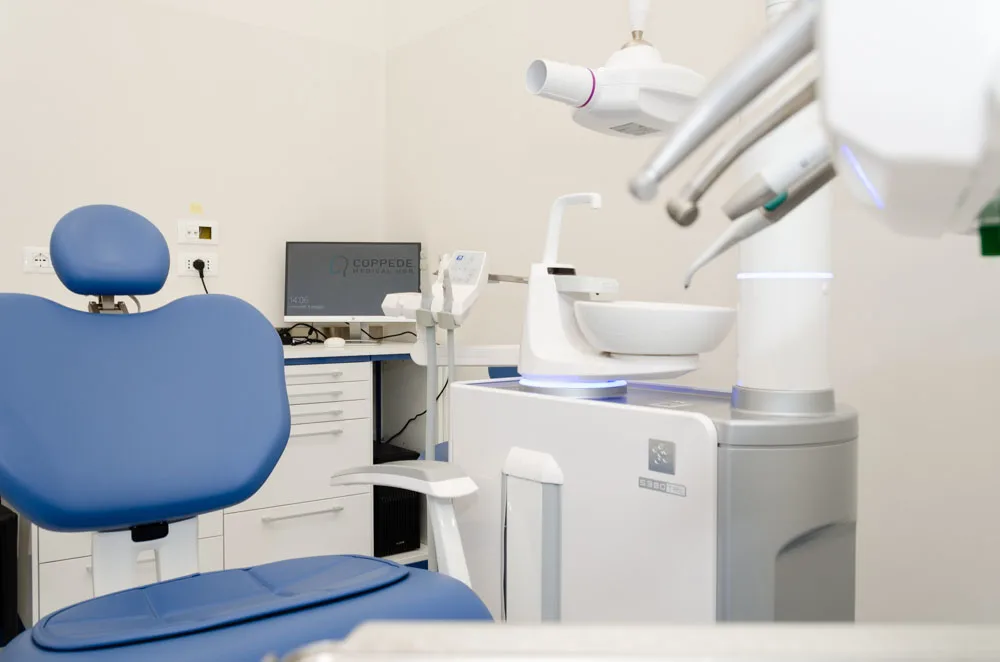 Un moderno studio dentistico presso lo studio-coppede-medical-hub-roma presenta una poltrona blu, attrezzature avanzate, un lavandino e un computer su un elegante mobile bianco.