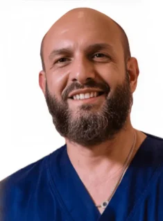 Un uomo sorridente con la testa calva e la barba folta, che indossa un camice da medico blu navy e una collana, è raffigurato su uno sfondo bianco semplice presso il Polo Medico Dott. Emiliano De Sanctis Coppedè di Roma.