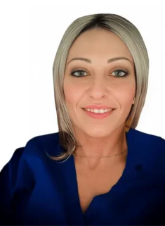 La Dott.ssa Flavia Aghilone Coppedè sorride all'obiettivo, con i suoi capelli biondi lunghi fino alle spalle e gli occhi azzurri completati da un top blu e una delicata collana, su uno sfondo bianco semplice al Medical Hub di Roma.