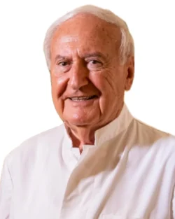 Il Dott. Arnold Brauner, un uomo anziano con i capelli bianchi, è sorridente e indossa una camicia bianca, in piedi davanti a uno sfondo bianco.