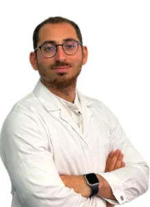 Il Dott. Claudio Saulli, un uomo con capelli corti, barba e occhiali, indossa un camice bianco e incrocia le braccia, in piedi su un semplice sfondo bianco.