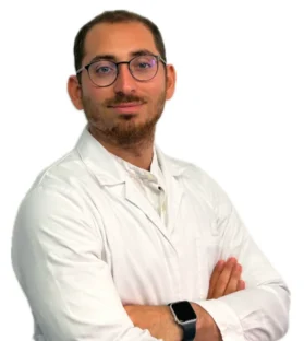 Il Dott. Claudio Saulli, un uomo con capelli corti, barba e occhiali, indossa un camice bianco e incrocia le braccia, in piedi su un semplice sfondo bianco.