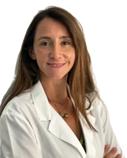 La Dott.ssa Claudia Milella Coppede, del Medical Hub Roma, con lunghi capelli castani e camice bianco, sta in piedi con le braccia incrociate e sorride alla telecamera su uno sfondo bianco.