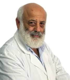 Uomo anziano con testa calva e barba bianca che indossa un camice bianco e una camicia azzurra, in posa su un semplice sfondo bianco presso il Centro Medico Dott. Guido Coen Coppede di Roma.